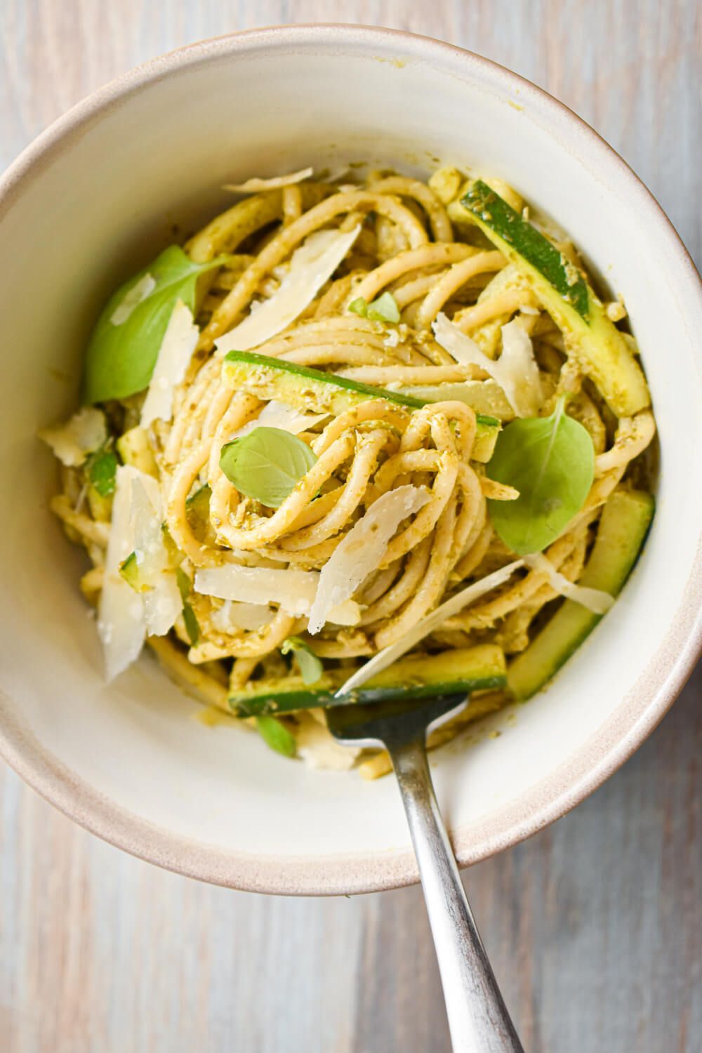 Pesto pasta with sliced zucchini, basil, and Parmesan cheese in a bowl.