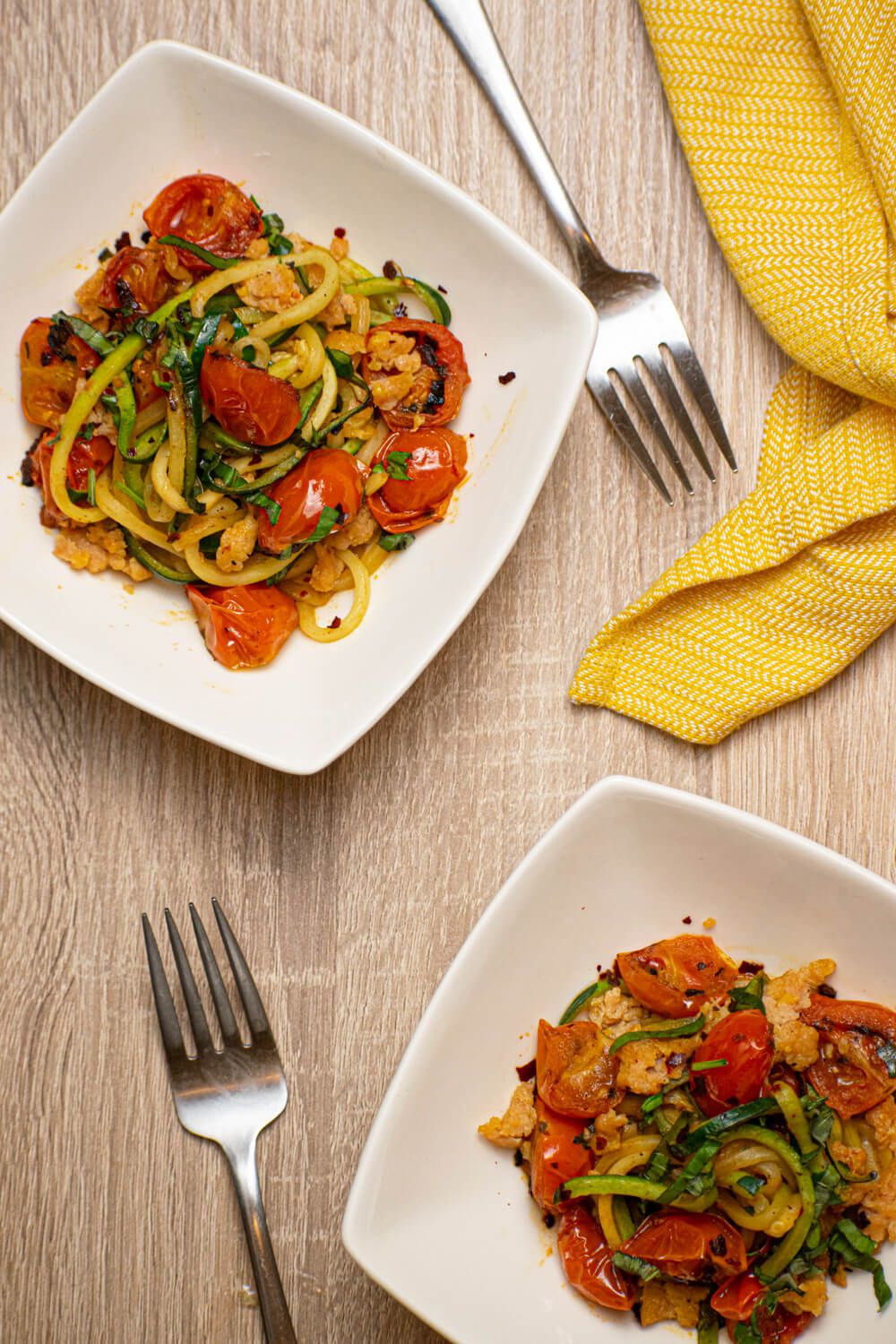 Zucchini noodles with sausage, cherry tomatoes, and garlic in two bowls with a yellow napkin.