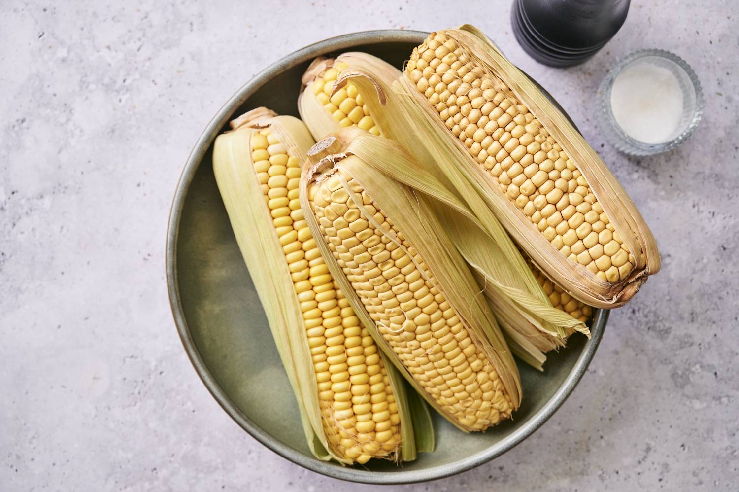 Fresh ears of corn in husks, arranged in a green bowl, with salt and a pepper grinder nearby on a light textured surface.
