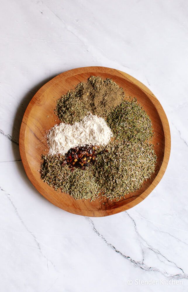 Italian spice blend with oregano, thyme, rosemary, basil, marjoram, garlic powder, and red pepper flakes on a wooden plate.
