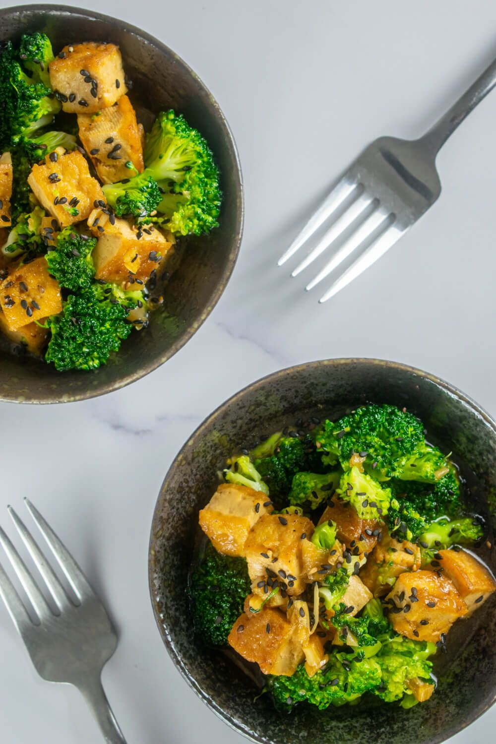 Sesame tofu with sesame seeds and broccoli in two black bowls with forks.