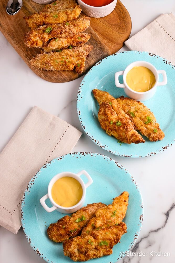 Crispy air fryer chicken tenders with honey mustard on two blue plates and a wooden cutting board.