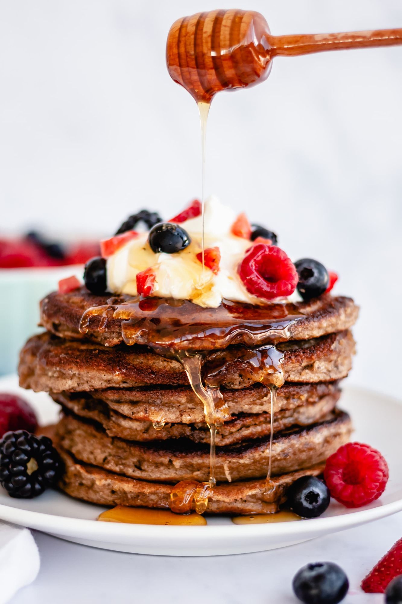 Almond flour and flax pancakes stacked on a plate with yogurt, berries, and maple syrup being drizzled on top.