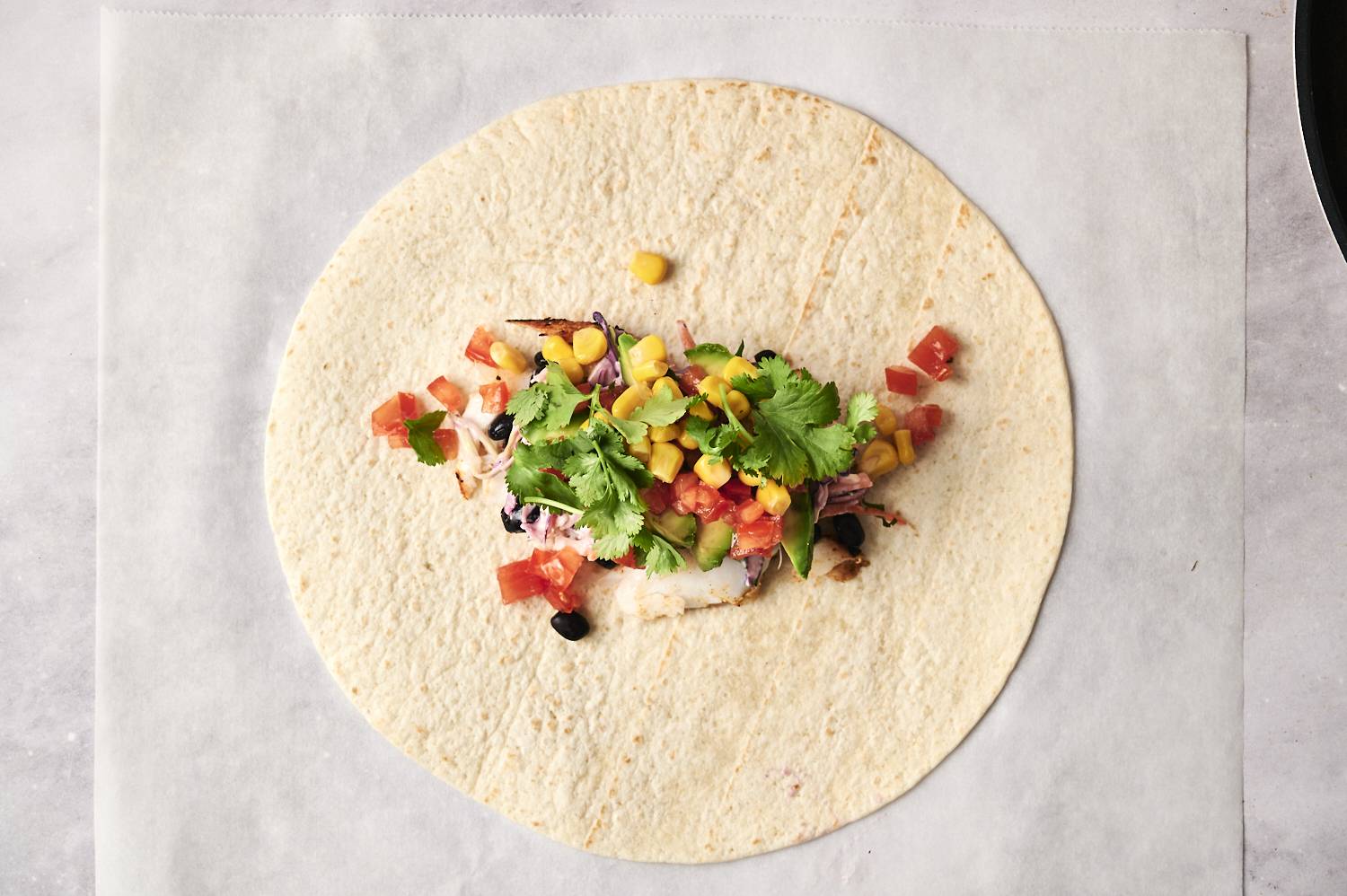 A tortilla with fish, coleslaw, black beans, corn, tomatoes, avocado, and cilantro, ready to be wrapped.