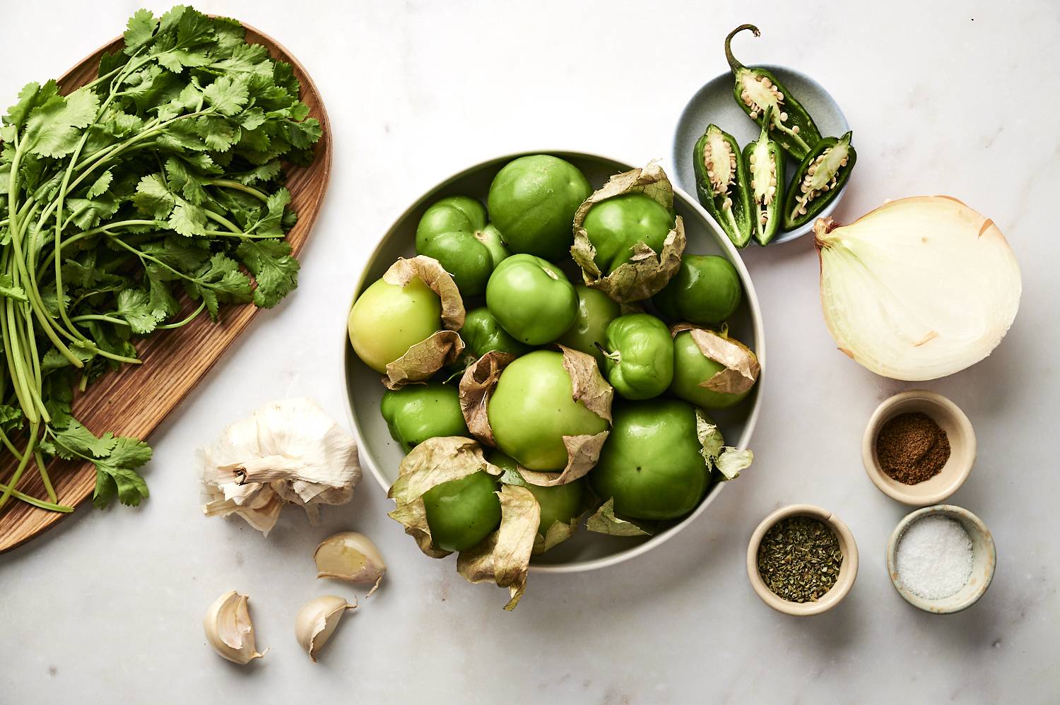 Ingredients for green enchilada sauce arranged on a white countertop, including fresh cilantro on a wooden board, a bowl of tomatillos with husks, sliced jalapeños on a small plate, garlic cloves, a halved onion, and small bowls of salt, dried oregano, and ground cumin.