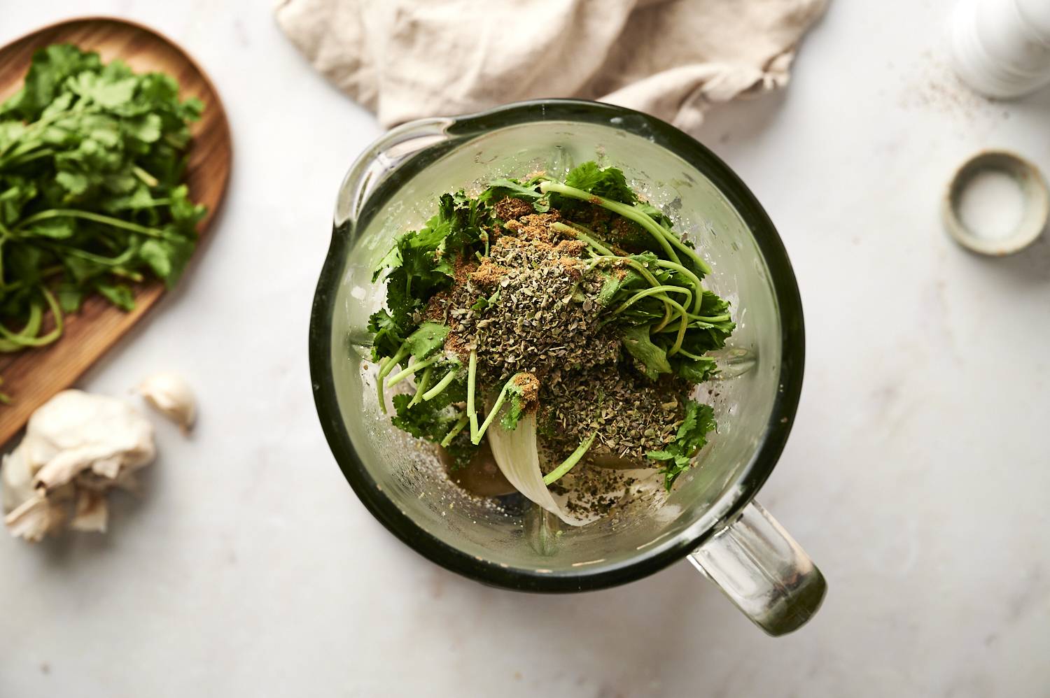 A blender jar filled with fresh cilantro, garlic, onion, tomatillos, and a mix of dried herbs and spices, ready for blending.