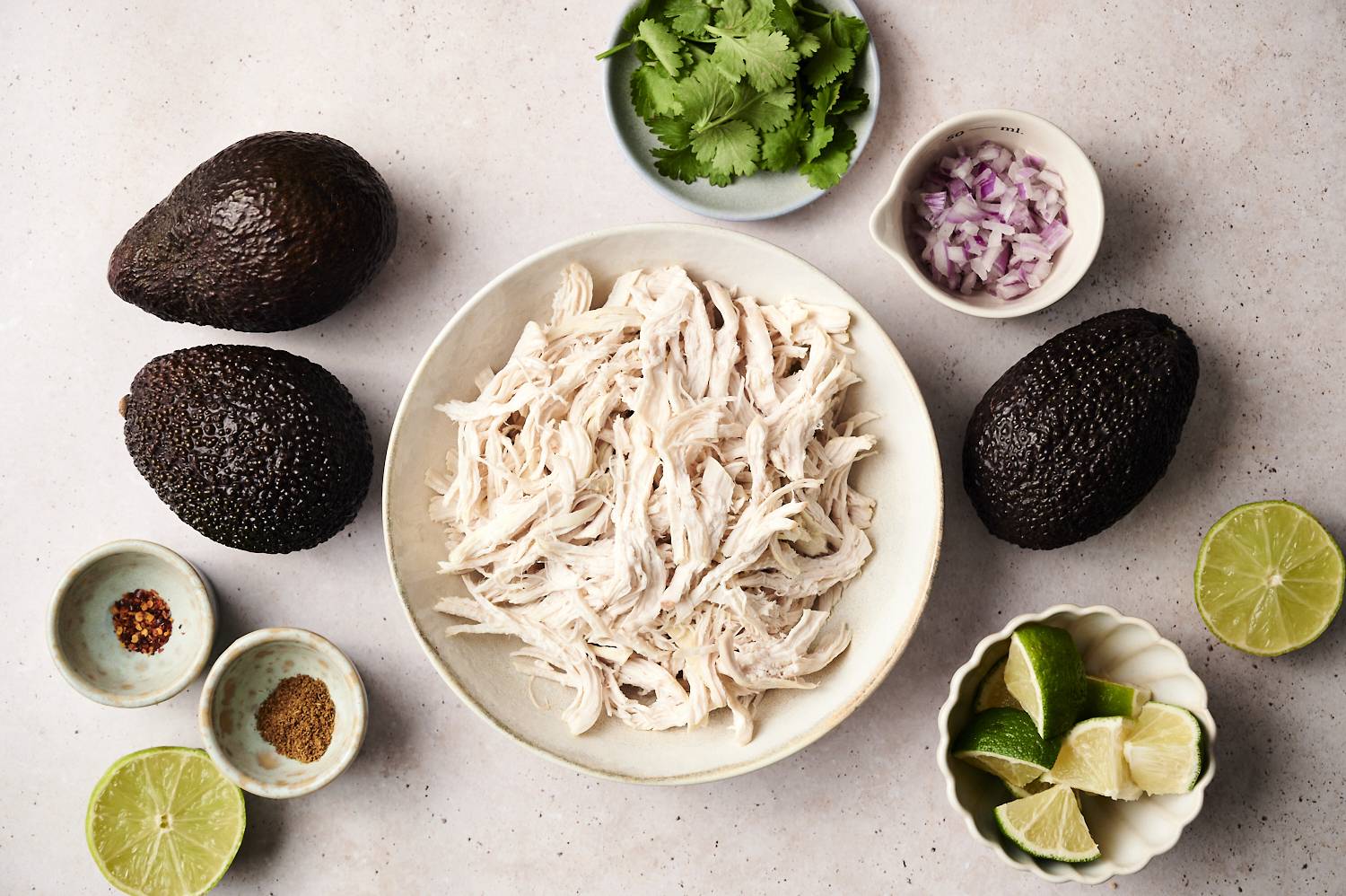 Flat lay of ingredients for avocado chicken salad, including a bowl of shredded chicken in the center, surrounded by whole avocados, chopped red onion, fresh cilantro, lime wedges, halved limes, ground spices, and red pepper flakes on a light countertop. 