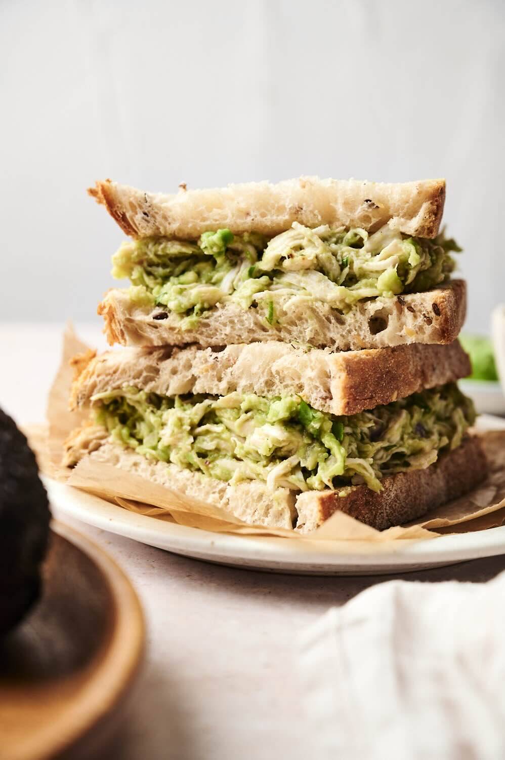 Close-up of a stacked sandwich made with creamy avocado chicken salad on thick slices of rustic bread. The filling is packed with shredded chicken and mashed avocado, served on a white plate with parchment paper.