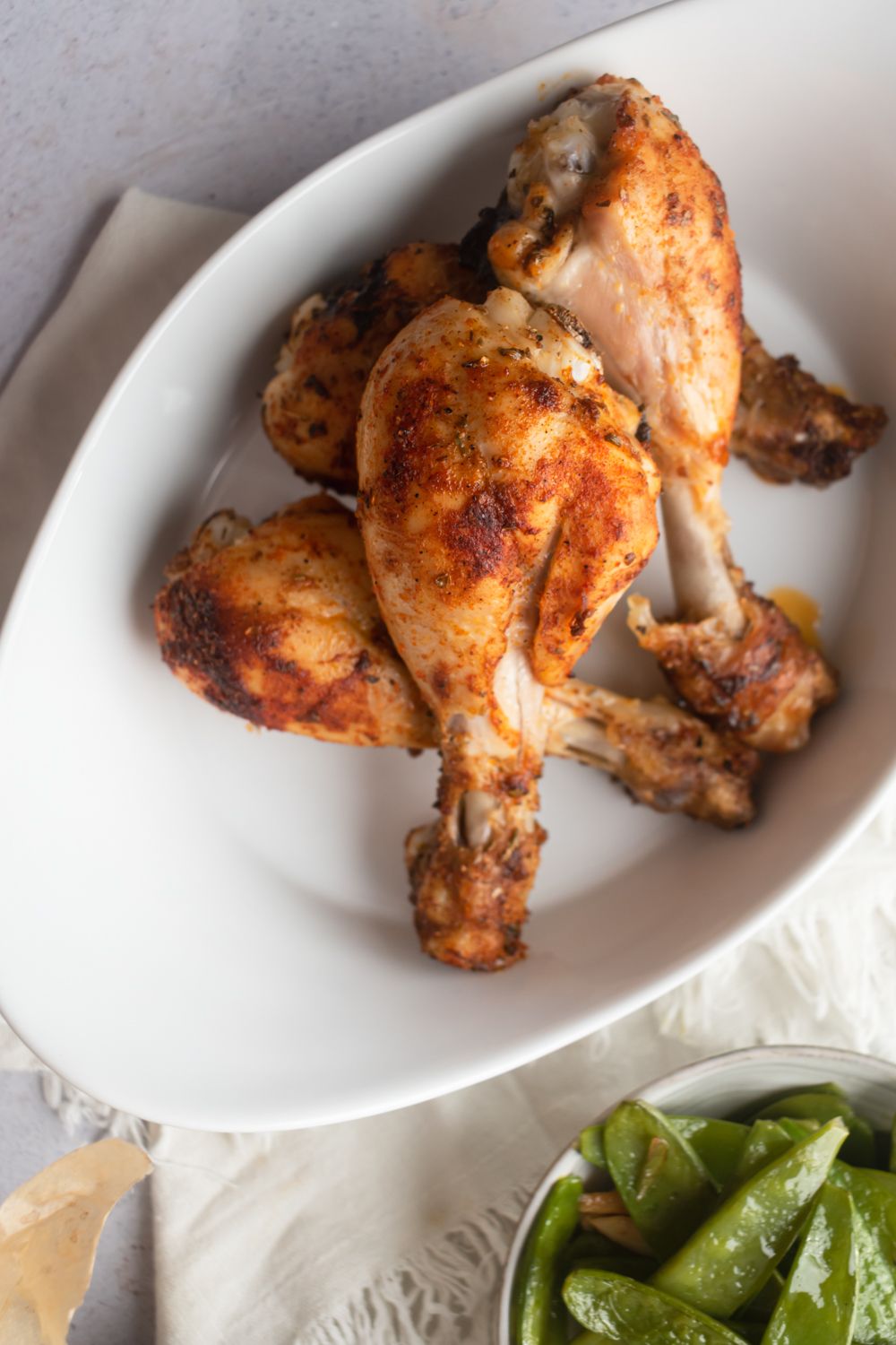 Skinless baked chicken drumsticks served in a white bowl with herbs and vegetables on the side.
