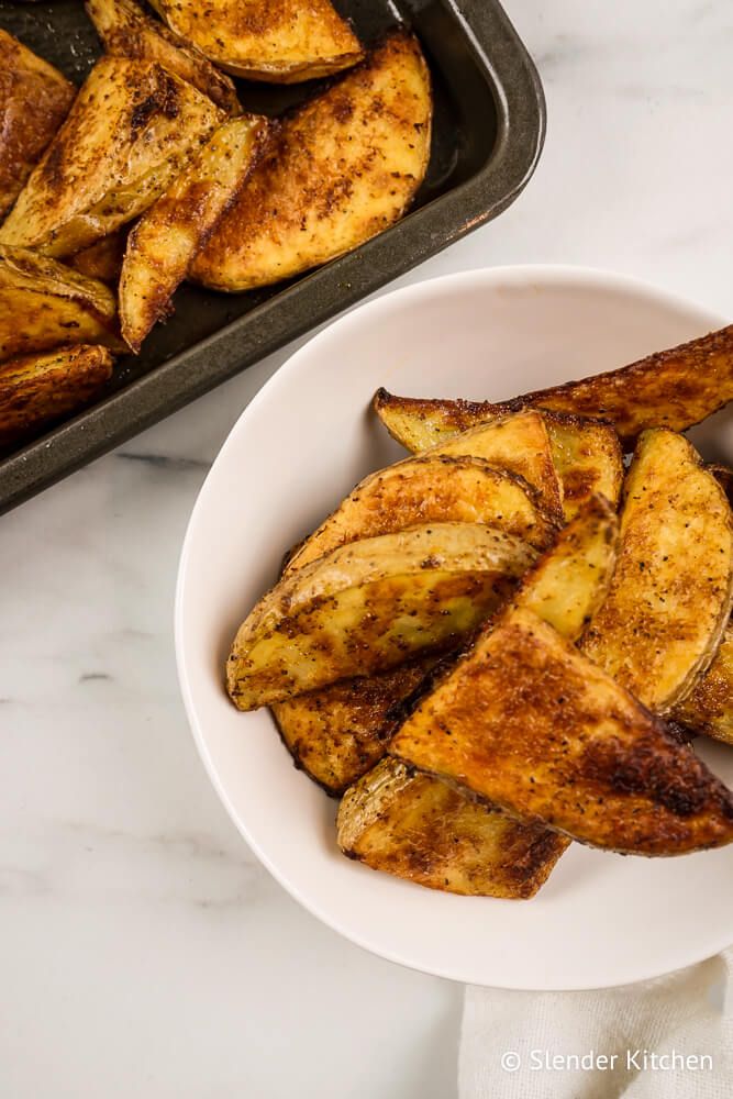 Potato wedges in a bowl and on a baking dish with garlic powder, paprika, salt, and pepper.