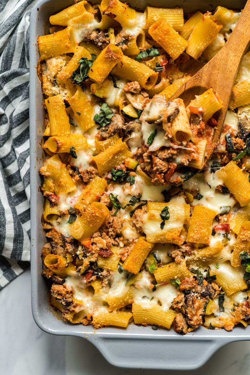 Baked bolognese ziti with zucchini, tomatoes, mushrooms, and melted mozzarella cheese in a dish with a wooden spoon.