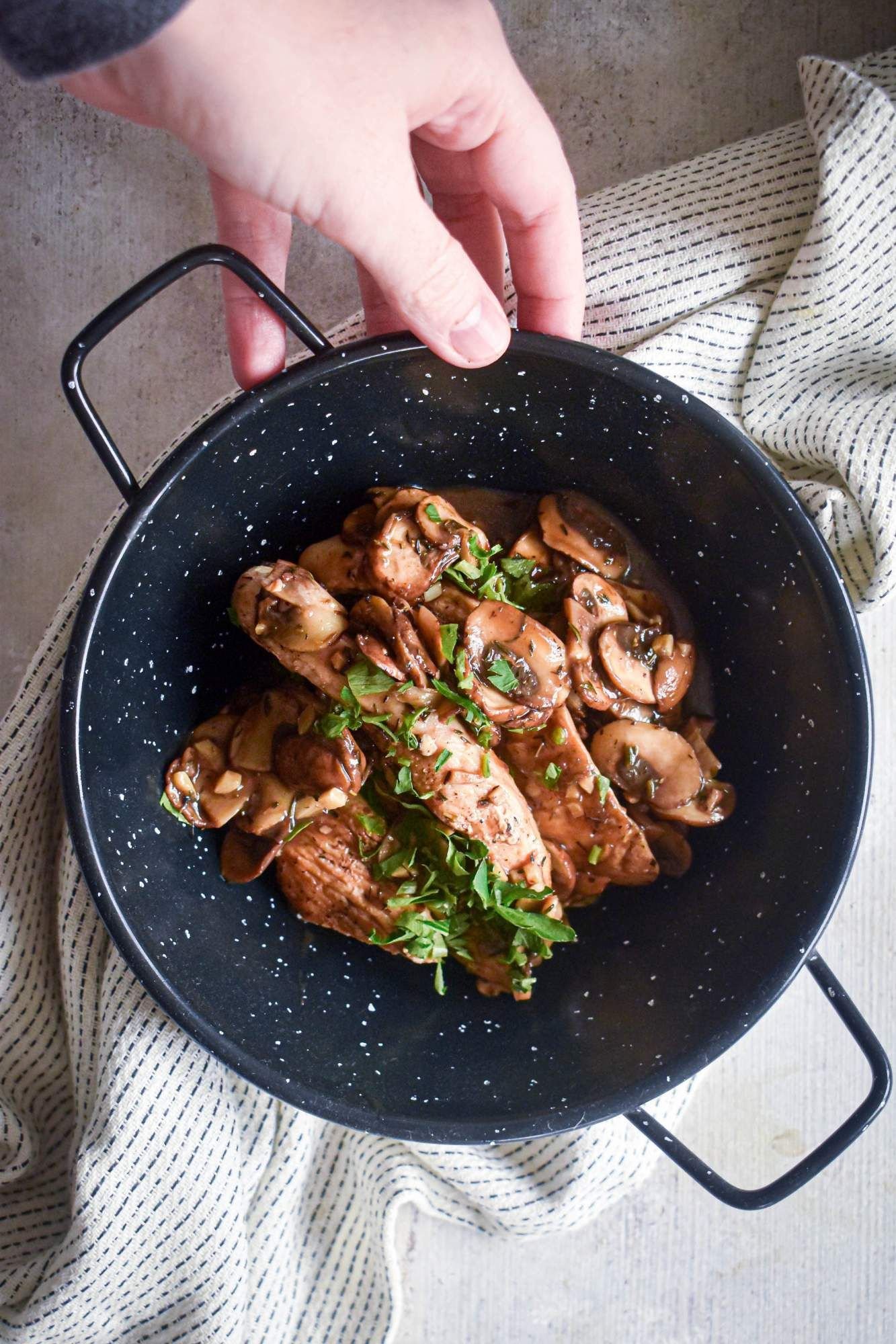 Balsamic chicken breasts with mushrooms and thyme in a blue bowl with a hand holding it.