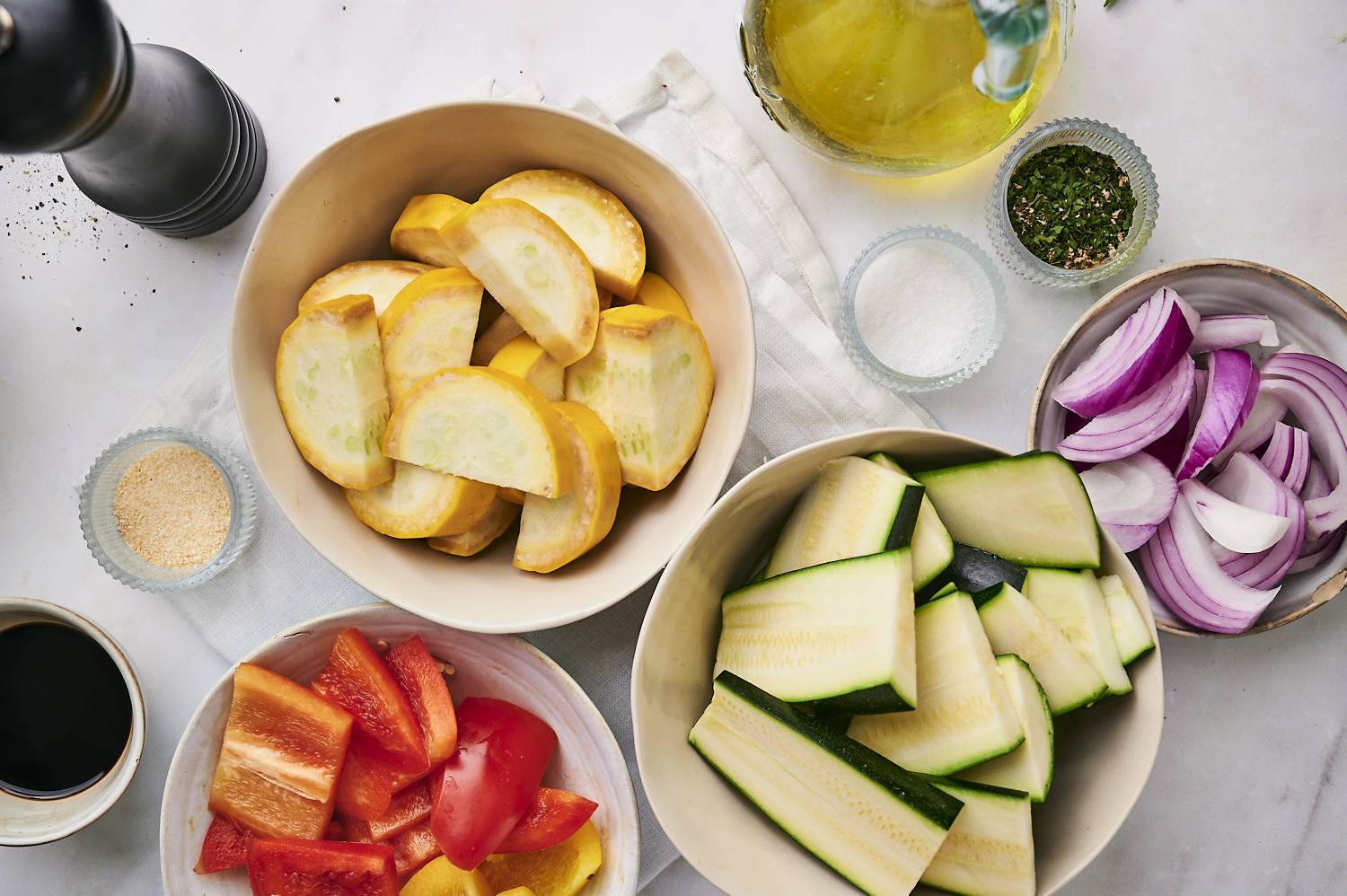 A countertop display of fresh vegetables: yellow squash, zucchini, red and yellow bell peppers, sliced red onion, and seasoning.