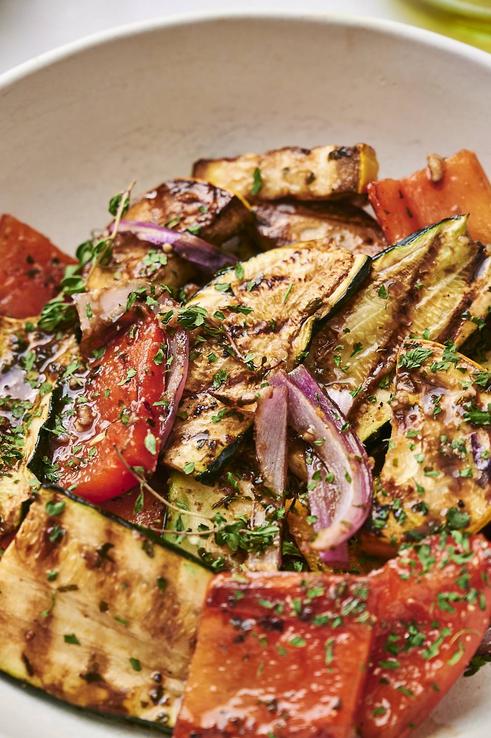 A close-up of a bowl filled with grilled zucchini, bell peppers, and onions, garnished with fresh herbs and a glossy dressing.