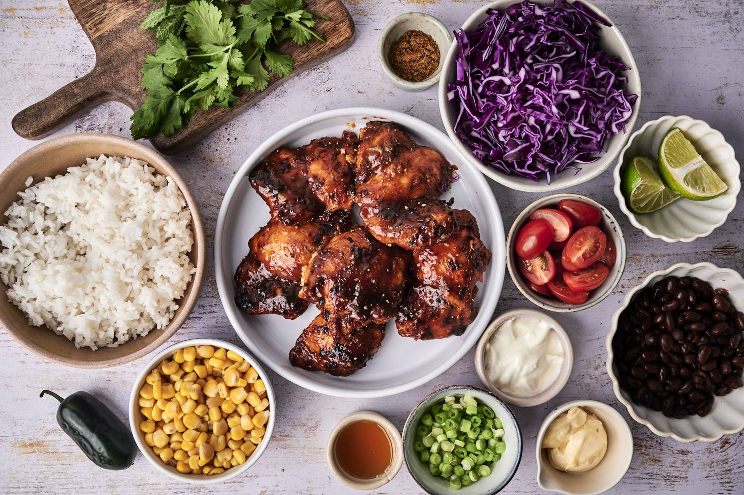 A colorful spread featuring grilled chicken thighs, rice, and various toppings: purple cabbage, cherry tomatoes, beans, corn, and lime.