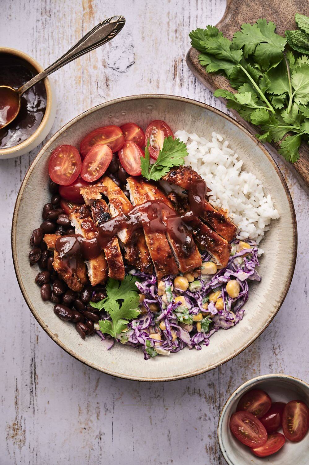 A vibrant bowl of chicken, rice, black beans, cherry tomatoes, and purple cabbage, garnished with cilantro and barbecue sauce.
