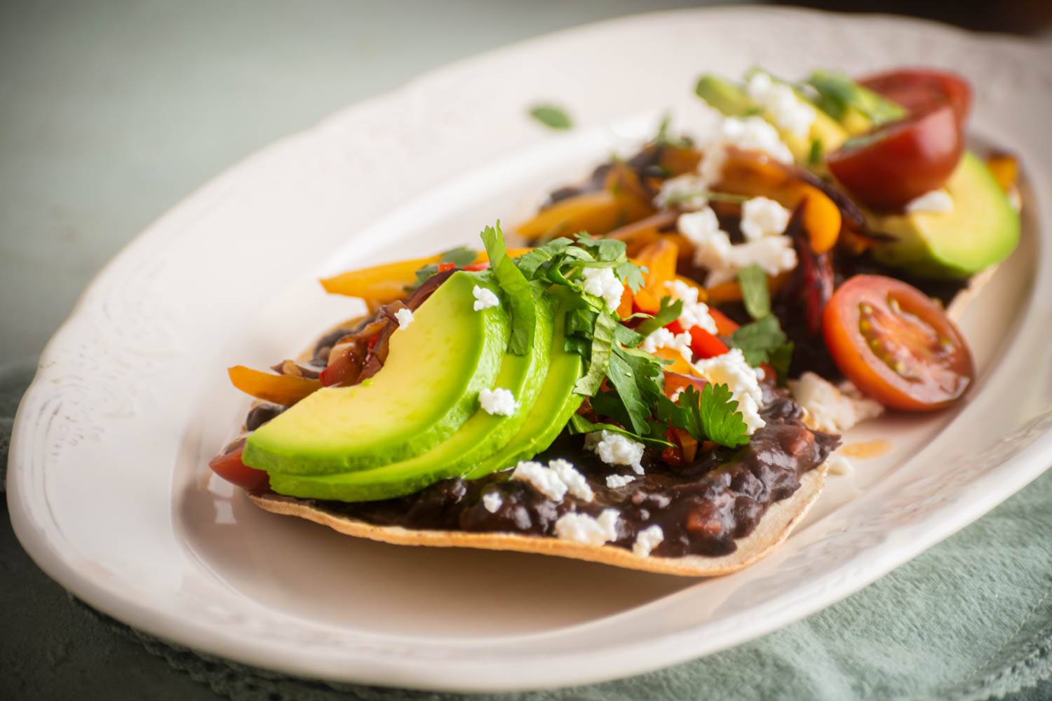 Close-up of a black bean tostada topped with sliced avocado, sautéed bell peppers, cherry tomatoes, crumbled cheese, and fresh cilantro on a white oval plate.
