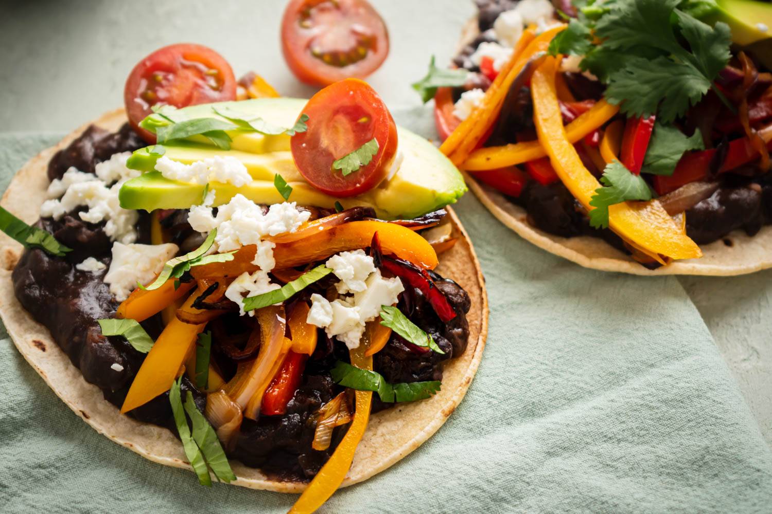 Two black bean tostadas topped with sautéed yellow and red bell peppers, sliced avocado, cherry tomatoes, crumbled cheese, and fresh herbs on a light green cloth.