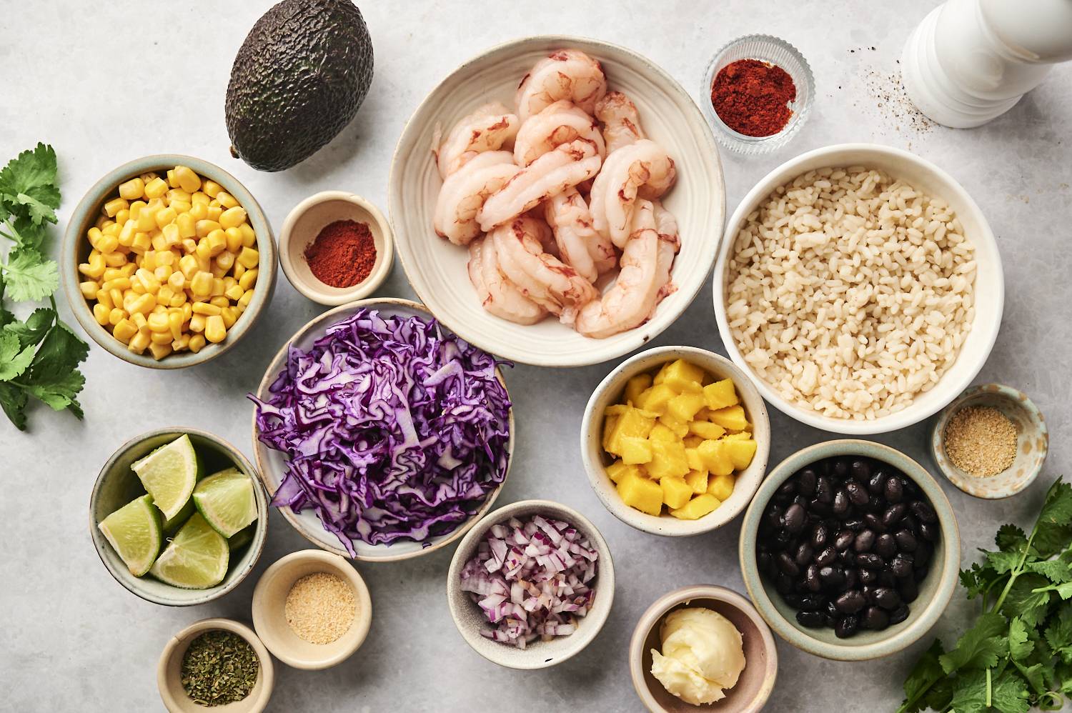 Ingredients for blackened shrimp bowls laid out, including shrimp, purple cabbage, mango, black beans, rice, corn, spices, and lime wedges.