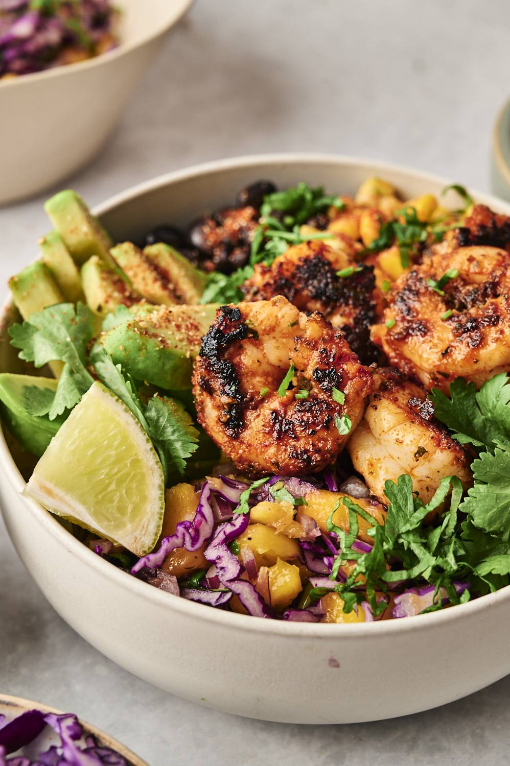 Close-up of a blackened shrimp bowl featuring charred shrimp, avocado slices, purple cabbage, mango, fresh cilantro, and a lime wedge.
