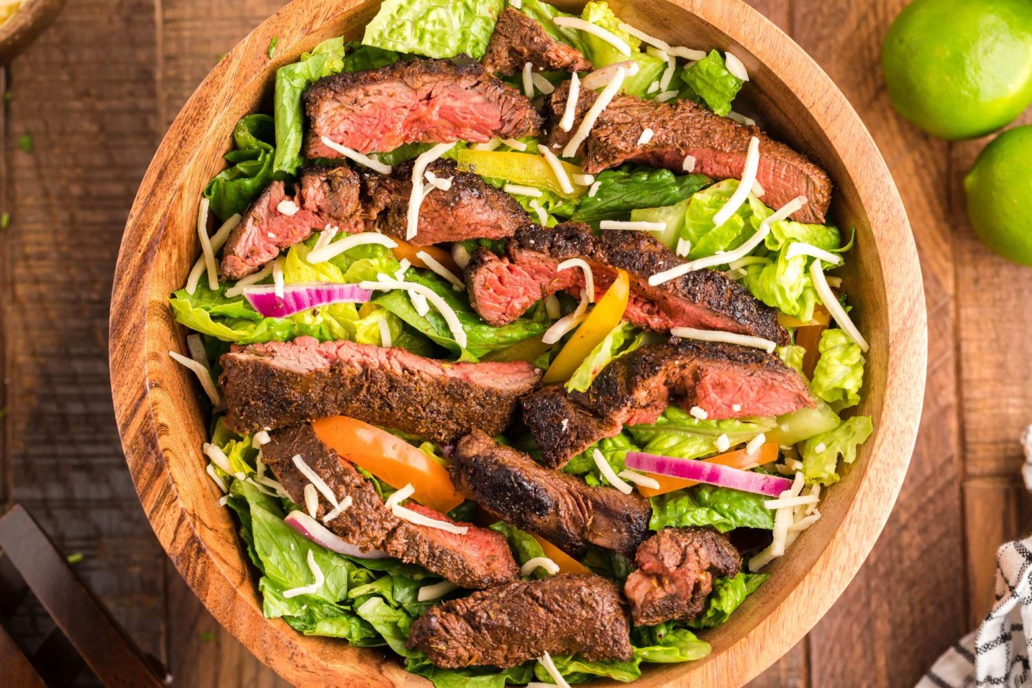 Steak salad with thinly sliced blackened steak in a bowl with lettuce, bell pepper, shredded cheese, and onion.