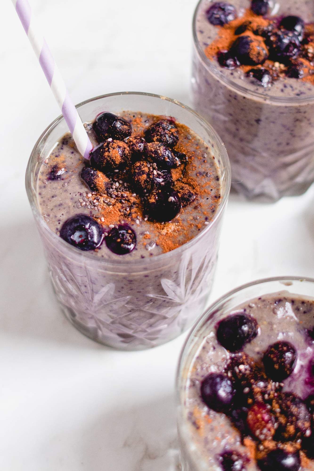 Banana and blueberry smoothies in a glass with frozen blueberries and cinnamon on top with a striped straw.