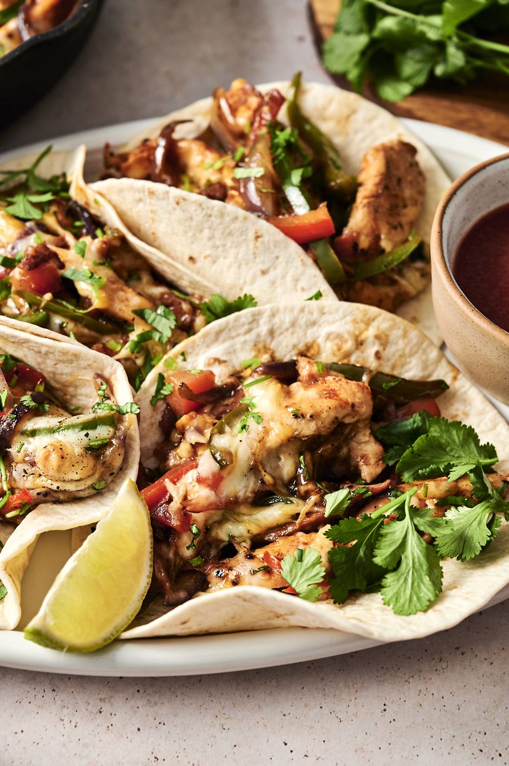 A close-up of chicken alambre tacos topped with melted cheese, cilantro, and lime wedges, with a side of salsa in a small bowl.