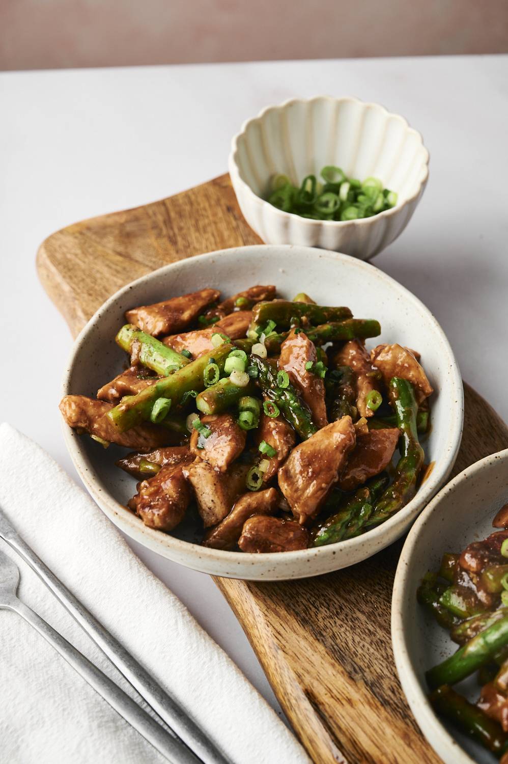 A bowl of stir-fried chicken and asparagus garnished with sliced green onions, served on a wooden board with a side bowl.