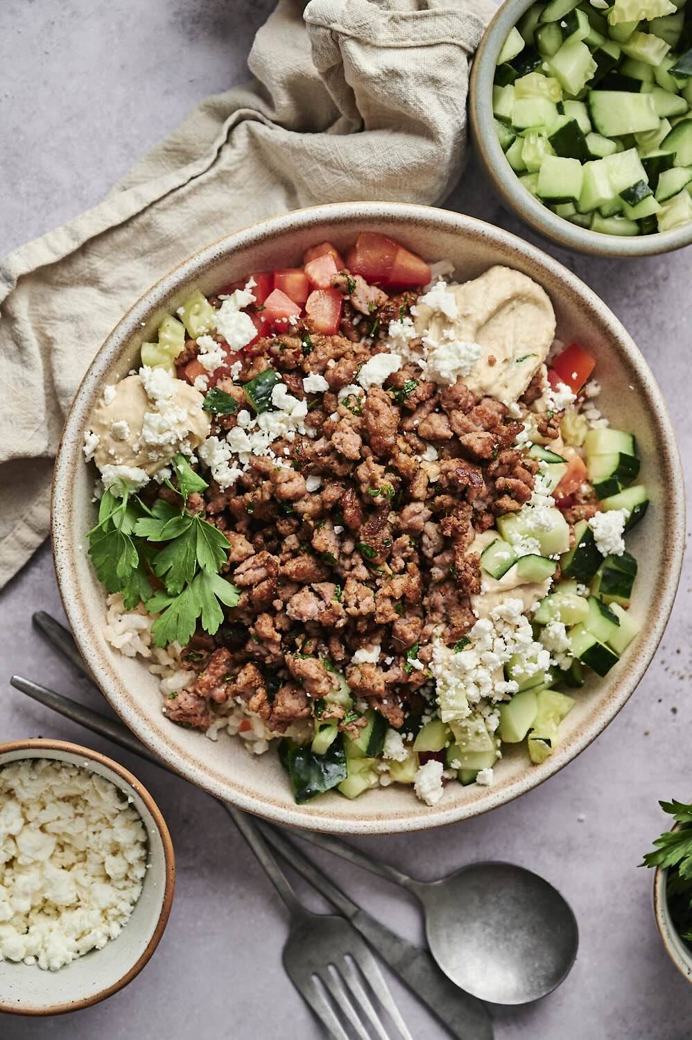 A vibrant bowl with spiced meat, fresh tomatoes, cucumbers, hummus, rice and feta, topped with cilantro.