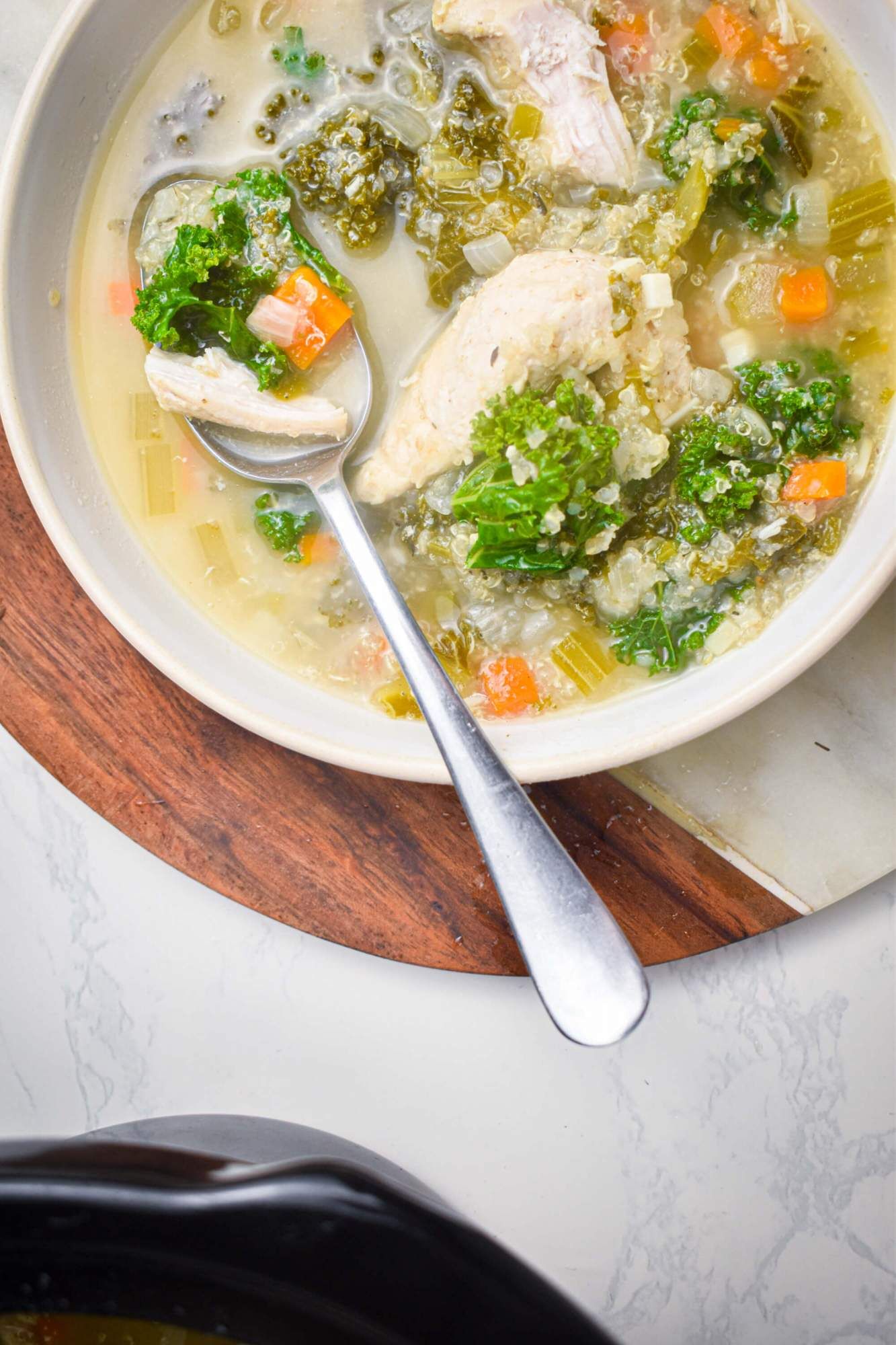 Chicken quinoa soup in a bowl with curly kale, chicken, vegetables, and broth in a bowl with a spoon.