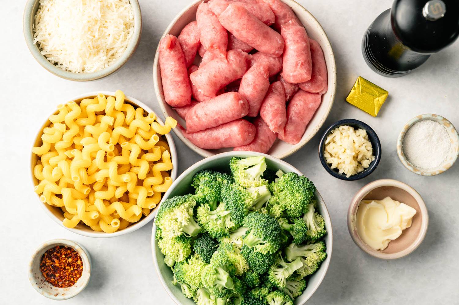 Bowls of raw sausage, broccoli florets, cavatappi pasta, grated cheese, minced garlic, and spices on a countertop. A pepper mill and butter are nearby.
