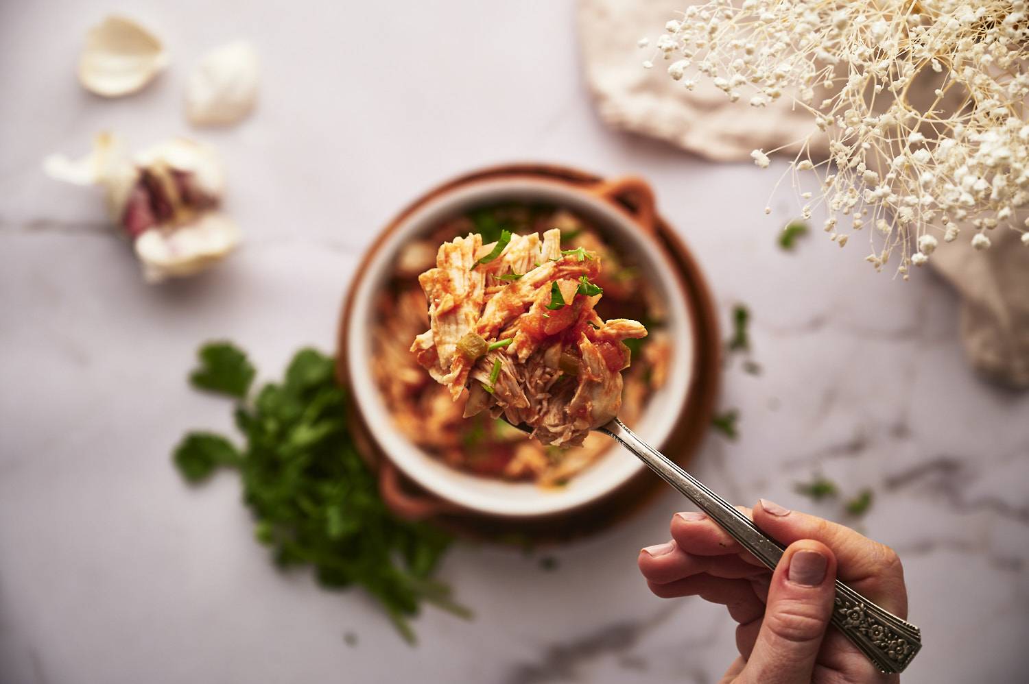 A hand holding a forkful of shredded chicken mixed with tomatoes and herbs, above a bowl on a marble surface with herbs nearby.