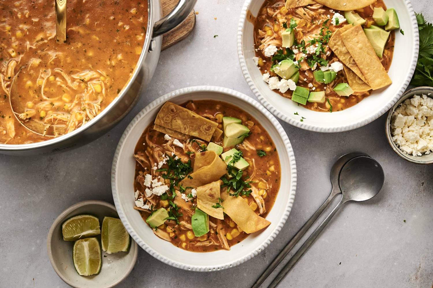 Top-down view of chicken tortilla soup served in white bowls, garnished with crispy tortilla strips, avocado, cilantro, and crumbled cheese, with lime wedges and a pot of soup in the background.
