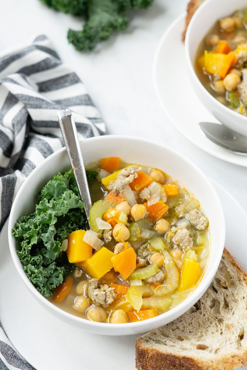 Chickpea and sausage stew with butternut squash in two bowls with kale and herbs on the side.