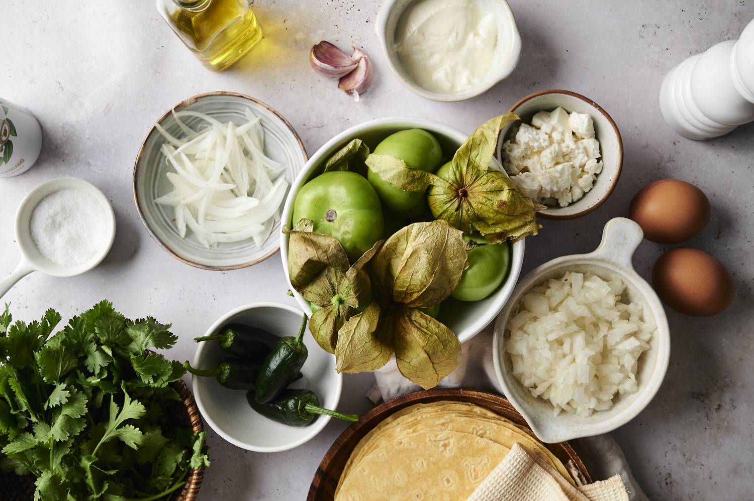 Fresh ingredients for chilaquiles verdes, including tomatillos, jalapeños, tortillas, onions, eggs, cheese, sour cream, and cilantro, arranged on a countertop.