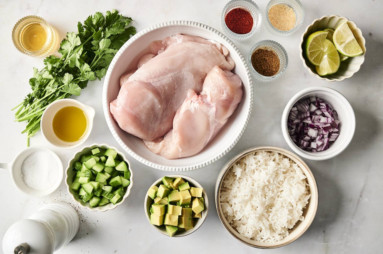 A flat lay of fresh ingredients including raw chicken, rice, avocado, cucumber, red onion, cilantro, lime wedges, and various spices, arranged on a white surface.