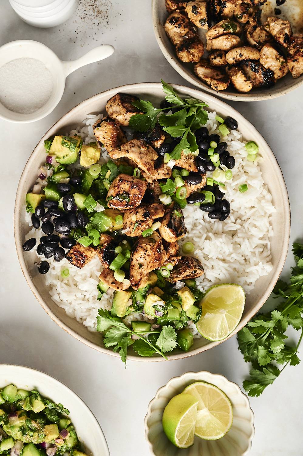 A close-up of a chili-lime chicken bowl with rice, black beans, avocado salsa, and fresh cilantro, garnished with lime wedges.