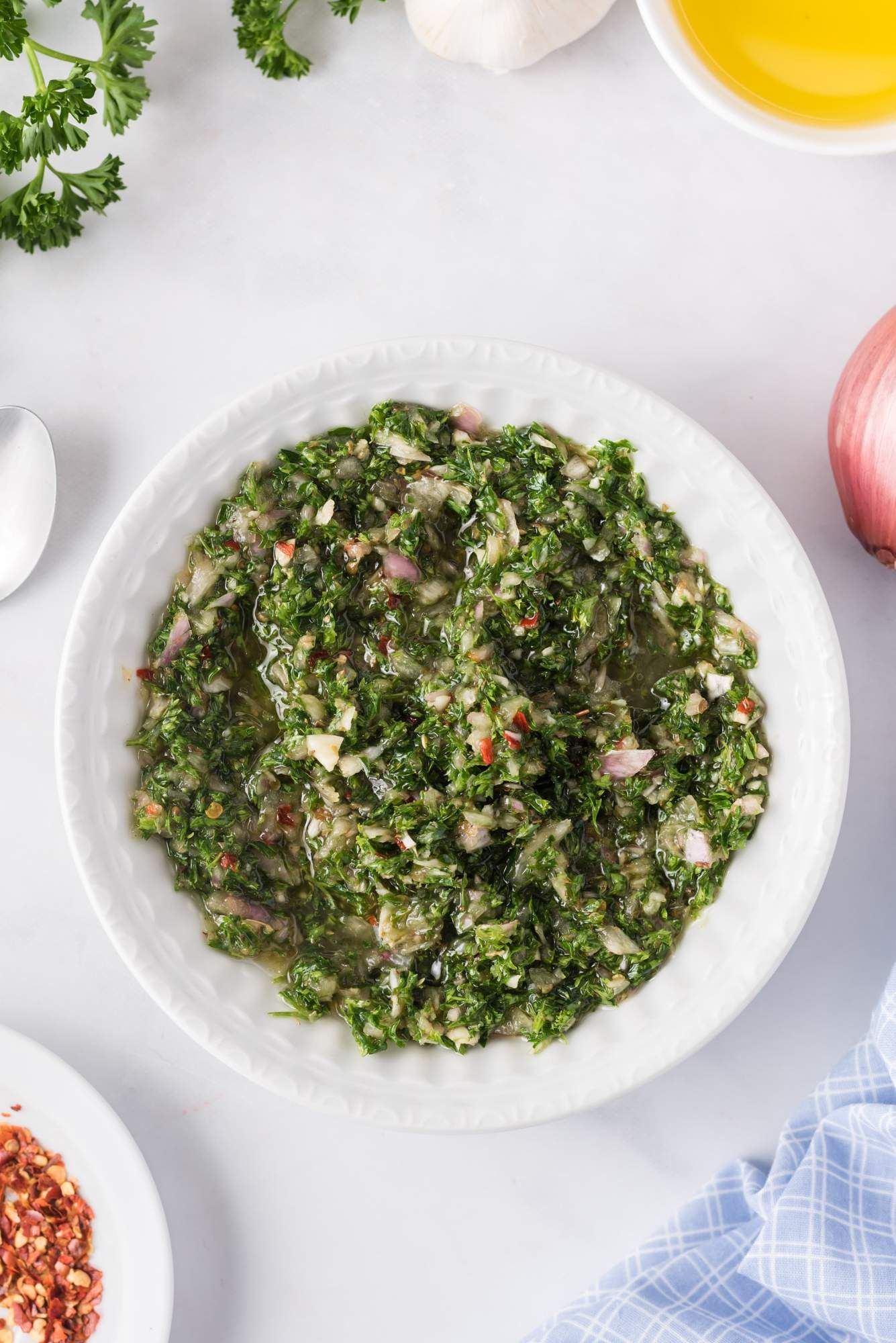 Chimichurri sauce served in a white bowl with parsley, shallots, and red pepper flakes on the side.