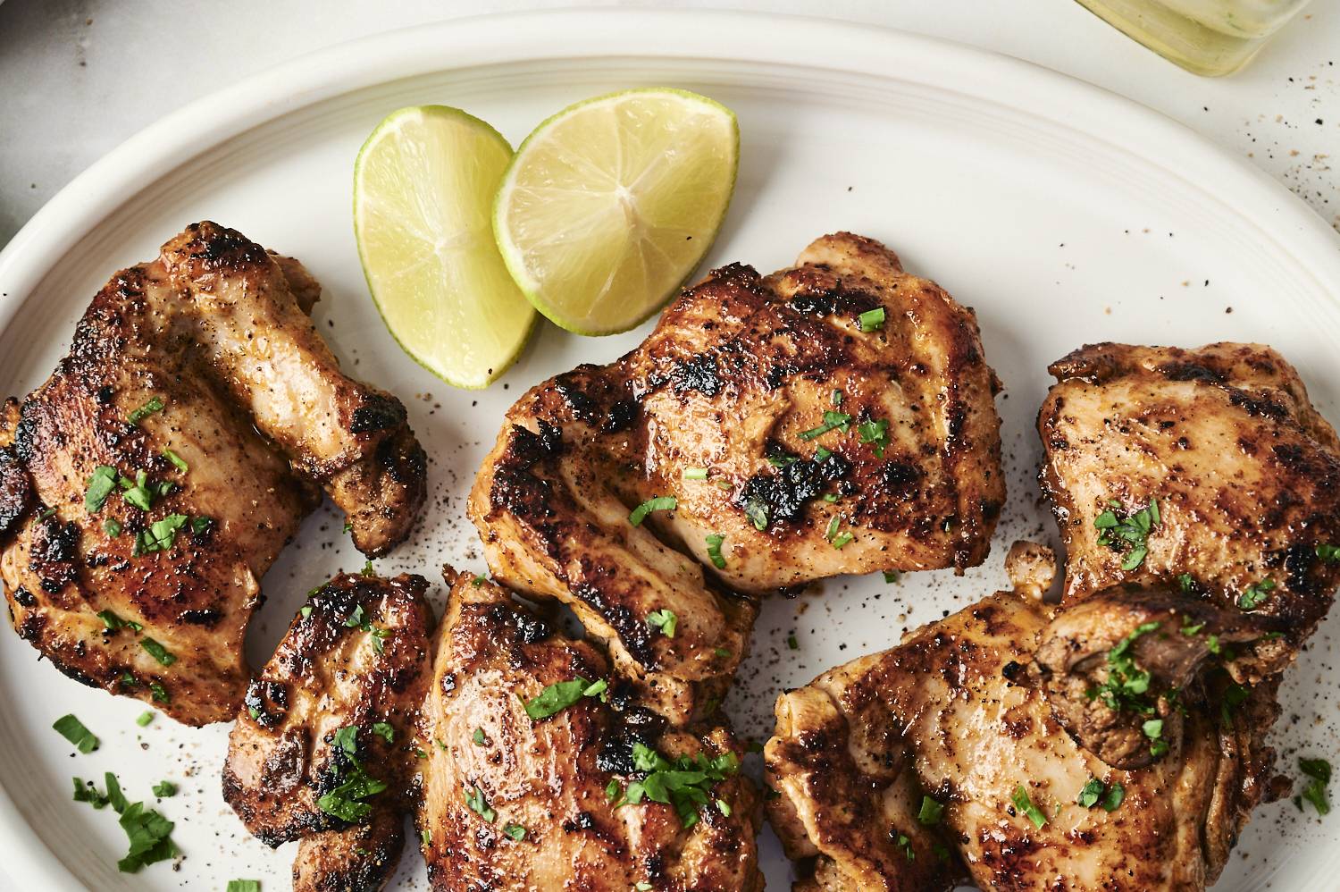 Grilled potato chip chicken thighs on a white plate decorated with fresh herbs and served with lemon wedges for a flavor.