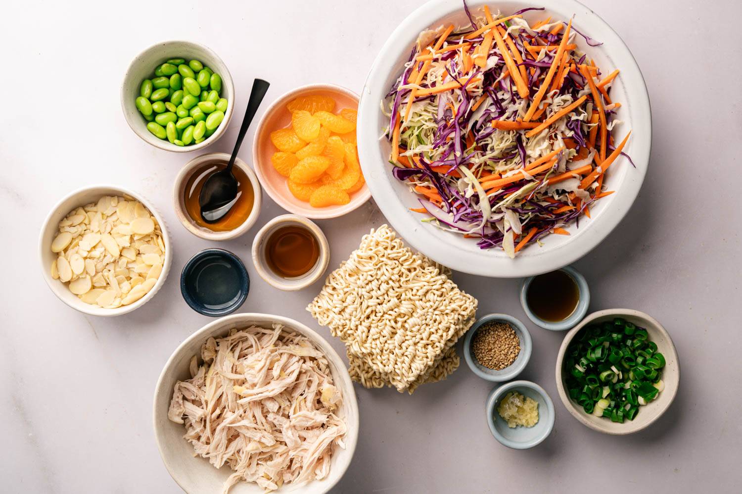 Ingredients for a vibrant Asian salad: shredded cabbage and carrots, edamame, mandarin slices, almonds, shredded chicken, ramen noodles, soy sauce, sesame seeds, green onions, and minced garlic on a light background.