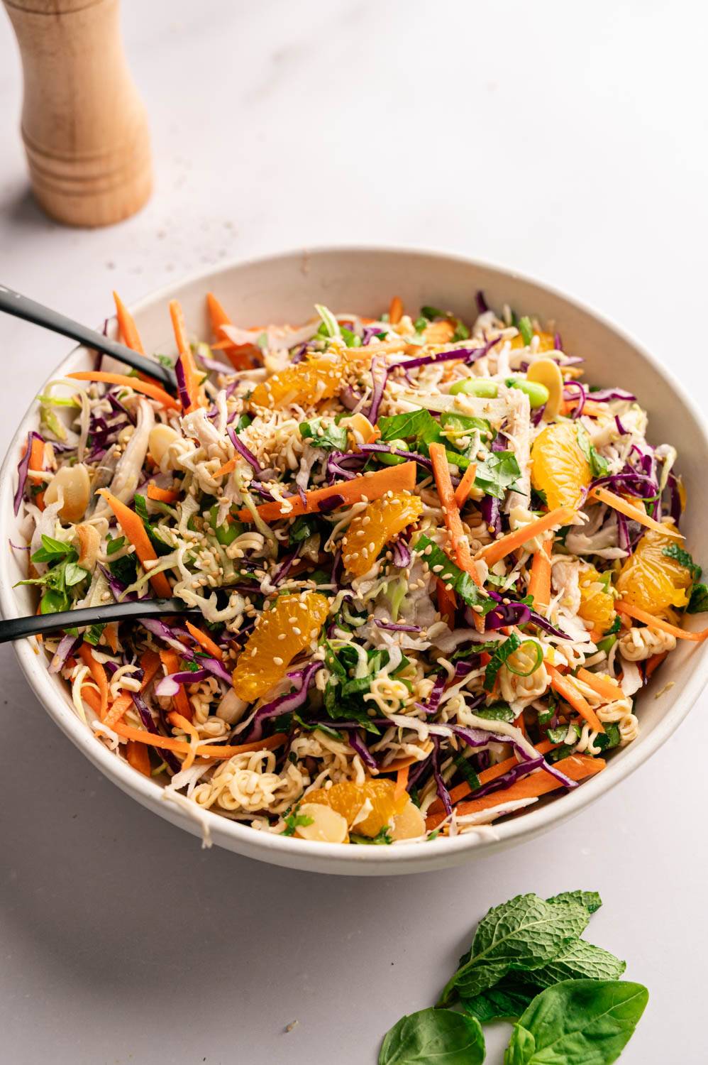A vibrant Asian salad in a bowl with vibrant purple cabbage, orange segments, and shredded carrots. Topped with sesame seeds and fresh herbs. Black chopsticks rest on the side.