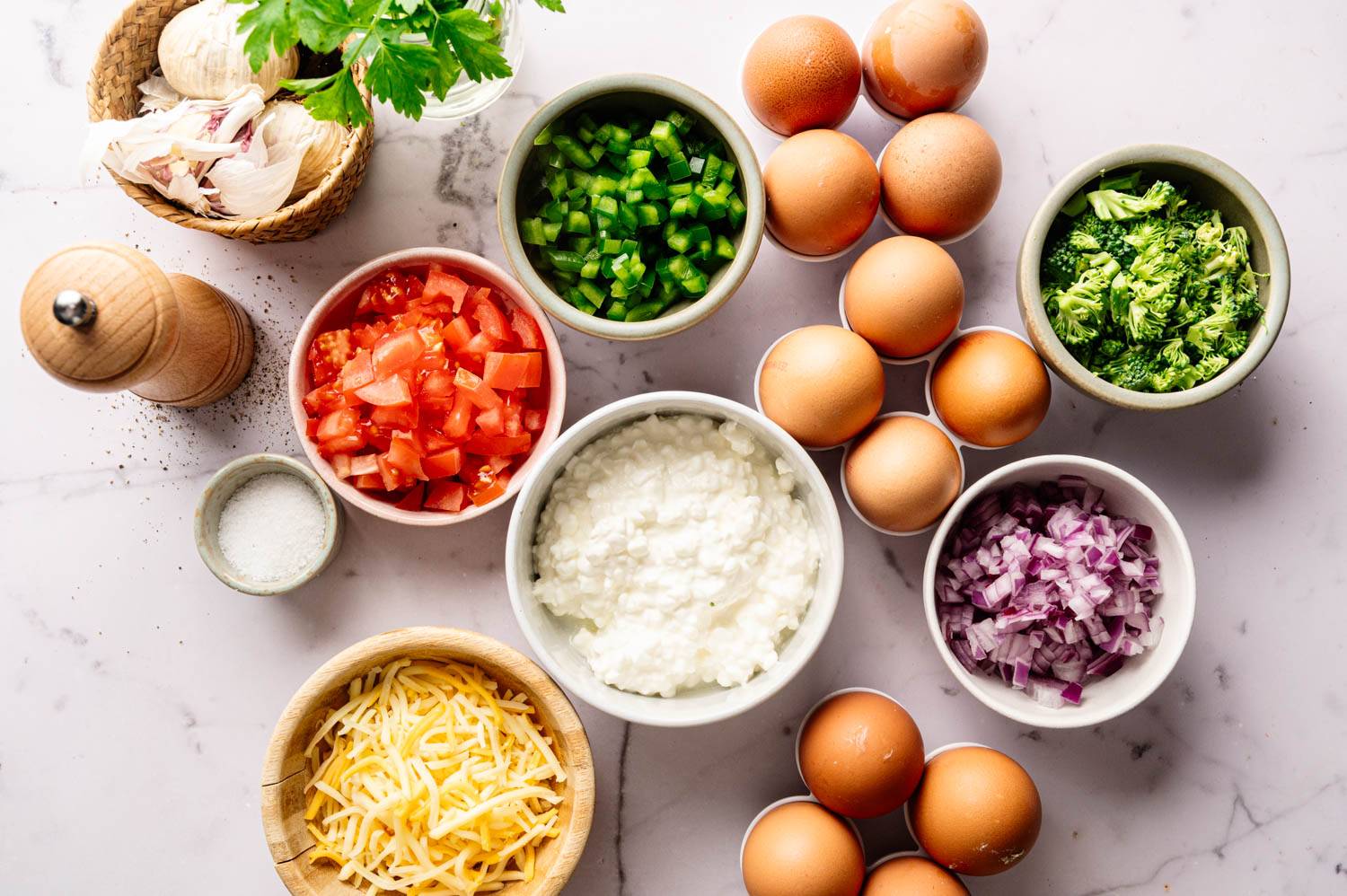 Ingredients on a marble surface: bowls of diced tomatoes, green peppers, red onions, broccoli, cottage cheese, shredded cheese, and a pile of eggs, surrounded by a basket with garlic, parsley, a salt bowl, and a pepper grinder.