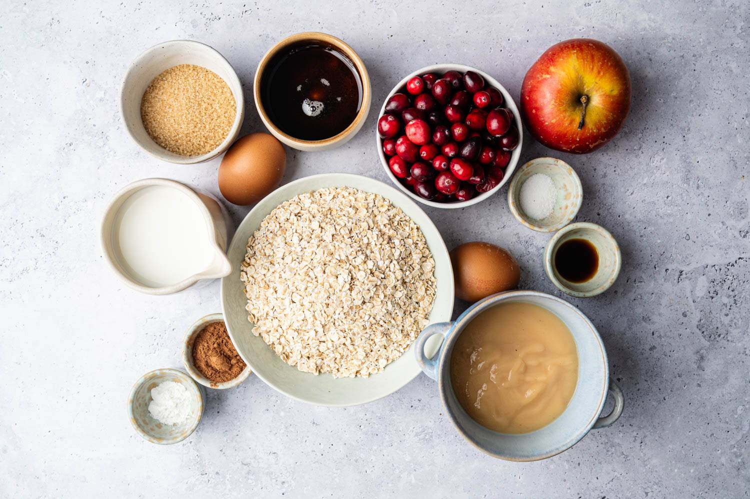 A collection of baking ingredients on a gray surface, including oats, cranberries, an apple, eggs, milk, brown sugar, vanilla, and sauce, arranged neatly.