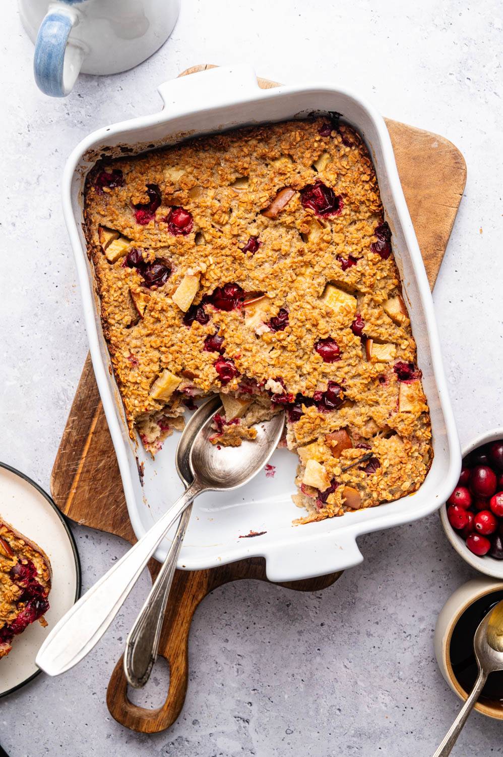 Baked oatmeal with apples and cranberries in a white rectangular dish on a wooden board. 