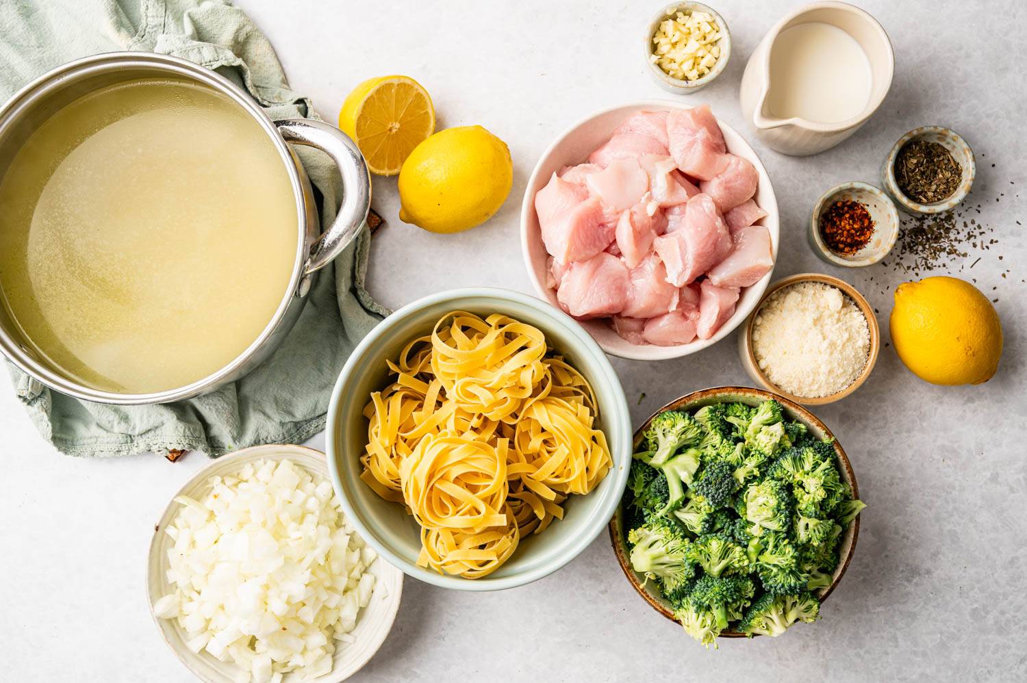 Various ingredients for a pasta dish are arranged on a countertop: raw chicken pieces, broccoli, tagliatelle, lemon, broth, onions, garlic, cream, Parmesan, and spices.