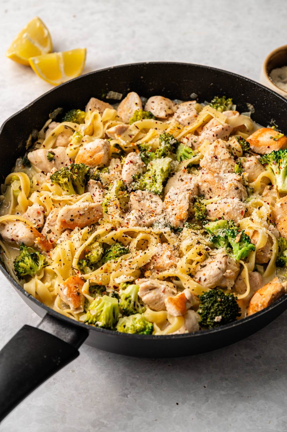 A skillet filled with creamy fettuccine Alfredo, broccoli florets, and tender chicken pieces, garnished with Parmesan cheese and pepper. Lemon wedges sit nearby.