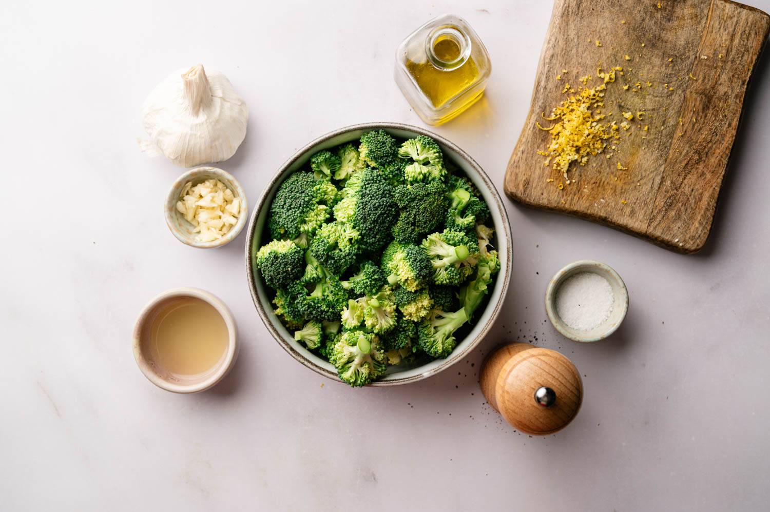 A bowl of fresh broccoli florets is surrounded by garlic, lemon zest on a cutting board, olive oil, salt, and a pepper grinder, creating a fresh, healthy vibe.
