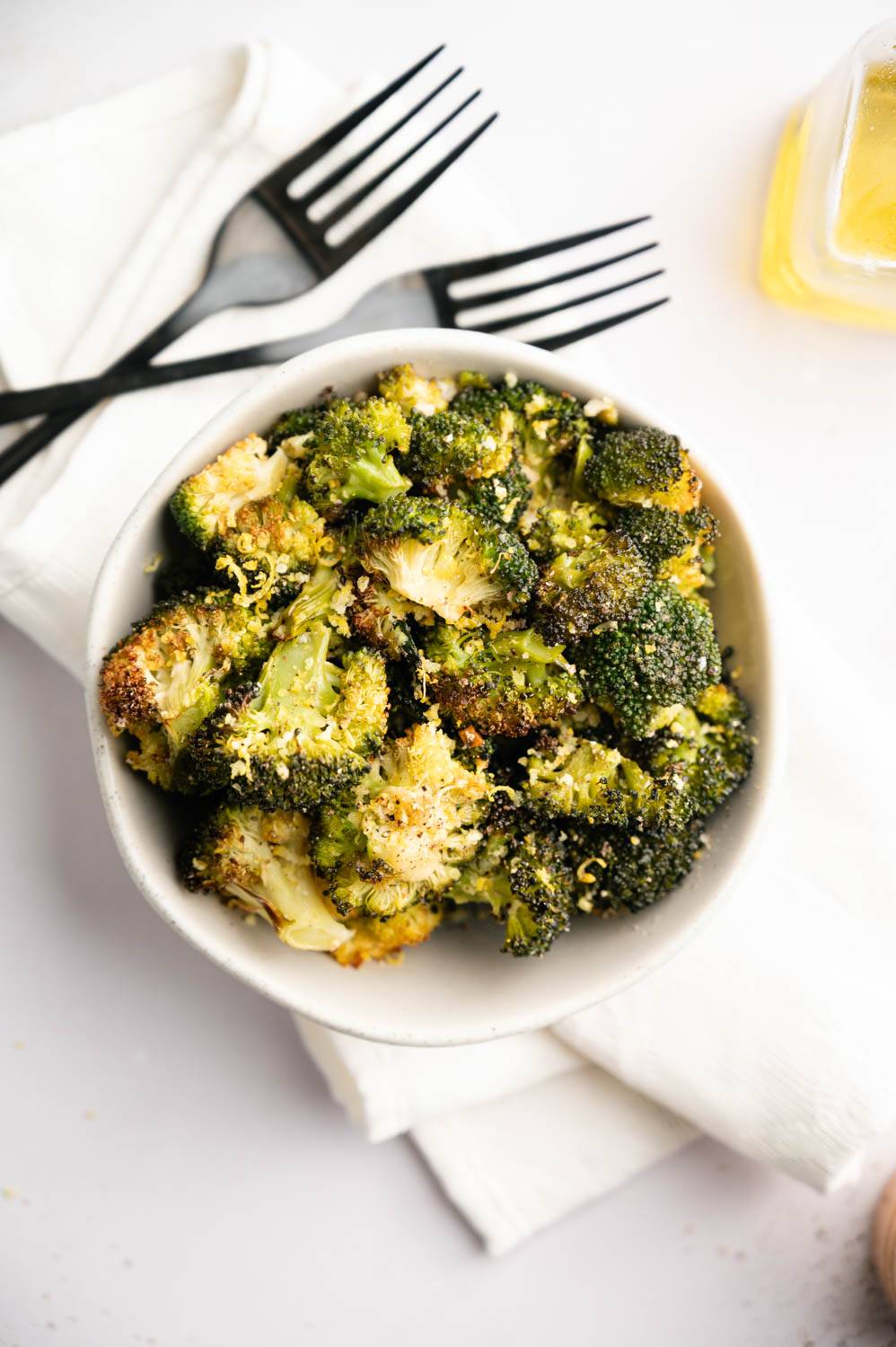 A bowl of roasted broccoli on a white cloth with two black forks. The broccoli is crisp and seasoned, exuding a warm, inviting aroma.