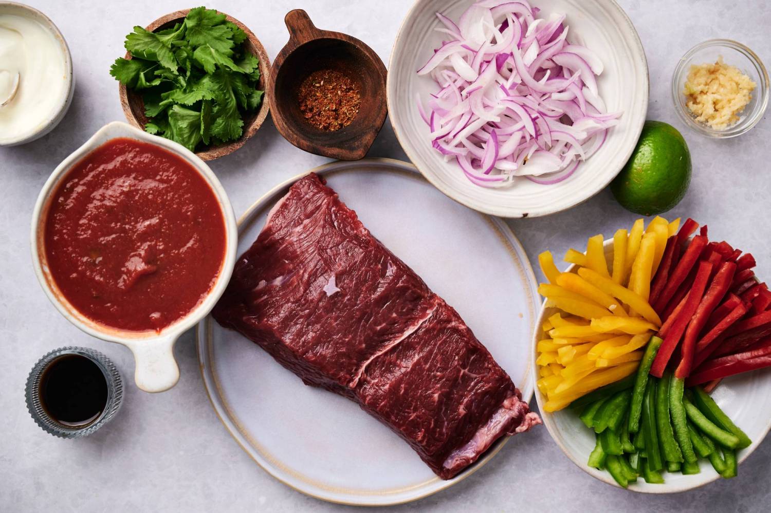 Ingredients for slow cooker beef fajitas including flank steak, bell peppers, onions, tomato sauce, garlic, cilantro, fajita seasoning, and limes.