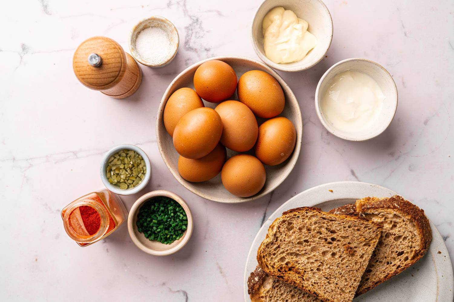 A collection of ingredients for egg salad: brown eggs, mayonnaise, yogurt, pickles, chives, paprika, salt, pepper, and slices of brown bread on a marble surface.